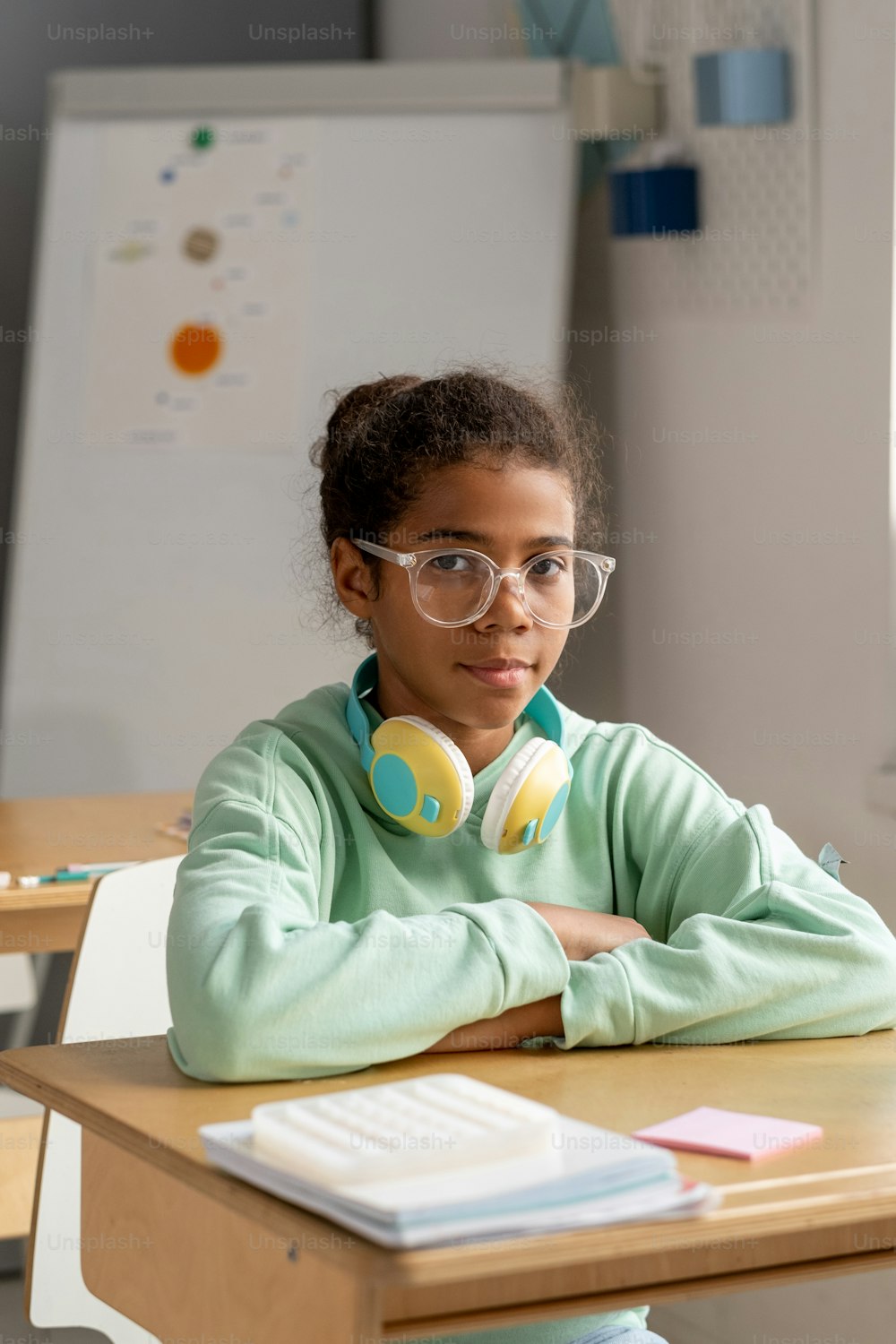 Jolie écolière sérieuse à lunettes assise près de son bureau et regardant la caméra à la leçon