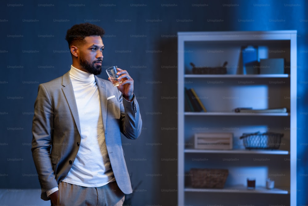 Un giovane uomo d'affari afroamericano con un bicchiere d'acqua in ufficio di notte, guardando di lato.