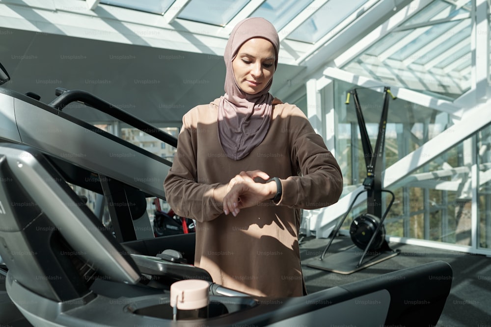 Ritratto medio orizzontale di una giovane donna musulmana adulta moderna che indossa l'hijab in piedi sul tapis roulant in palestra che controlla qualcosa sul suo tracker di attività