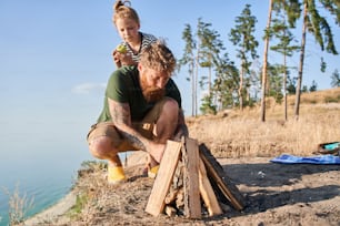 Incredible adventure. Little cute daughter eating apple near the future campfire. Her tattooed father preparing bonfire with his little helper