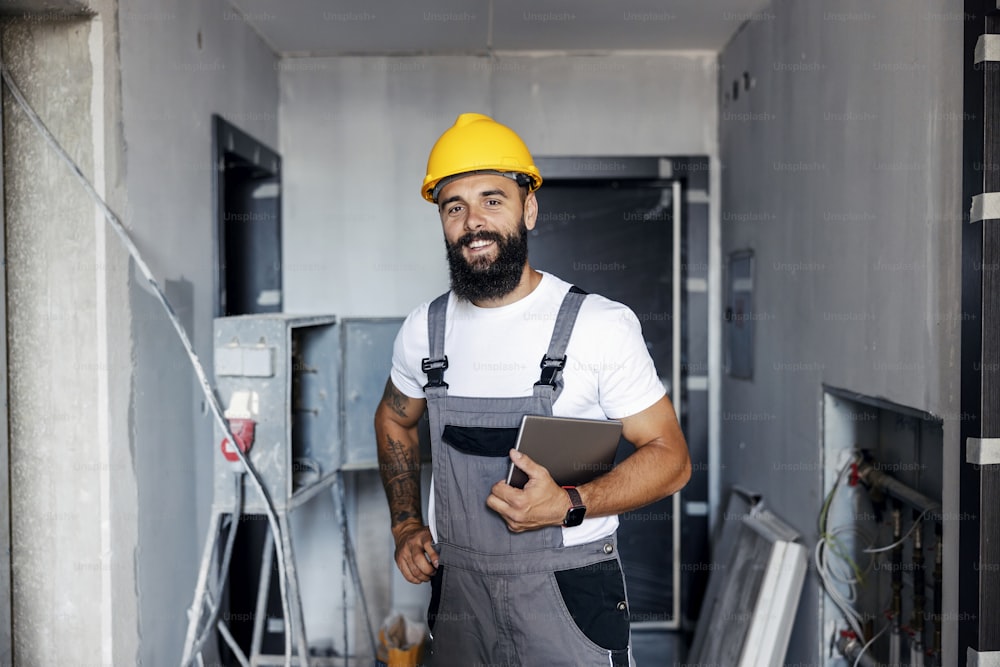 A happy construction worker with a helmet is standing in an unfinished building and holding a tablet under the armpit. He is checking on works on site.