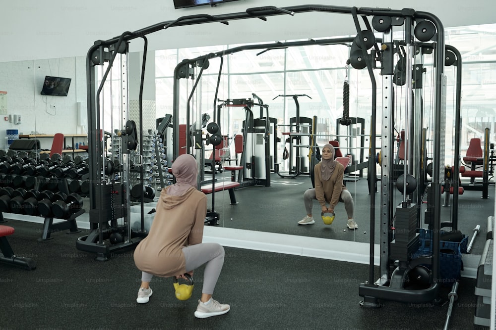 Horizontal long shot of sporty Muslim woman wearing headwrap doing squats exercise with kettlebell in front of mirror in gym