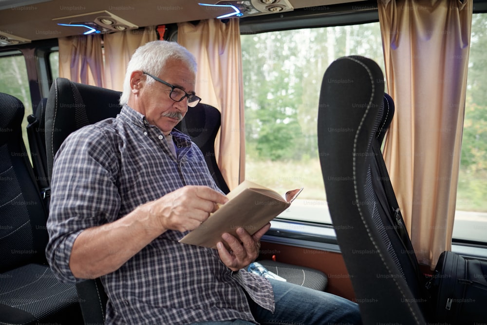 Homme caucasien âgé concentré avec des cheveux blancs lisant un livre tout en prenant un bus à travers la forêt