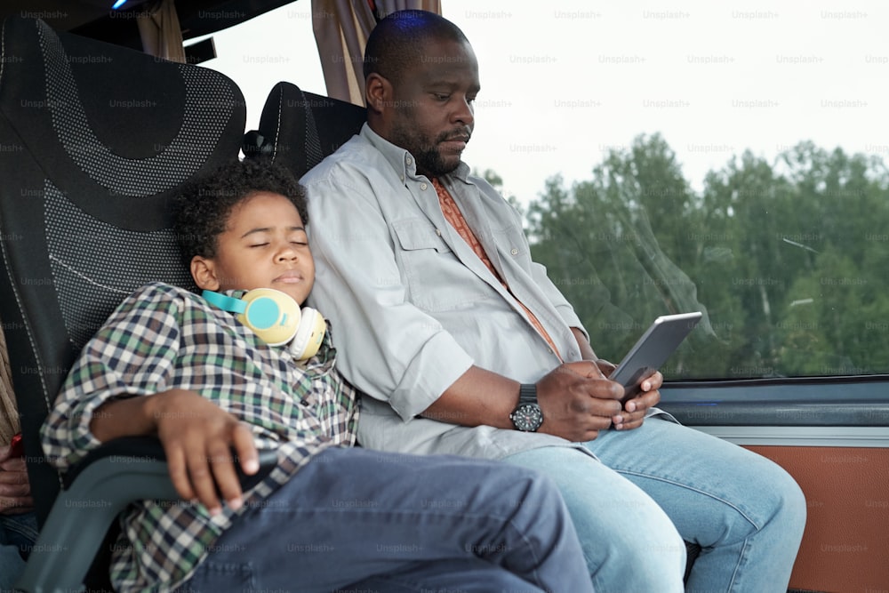 Afro American boy with headphones around neck sleeping on fathers shoulder in bus while he using tablet
