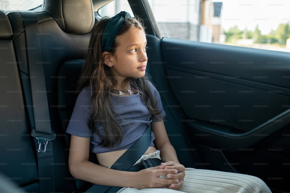 Bambina che si siede sul sedile posteriore della nuova auto elettrica confortevole e che guarda attraverso il finestrino