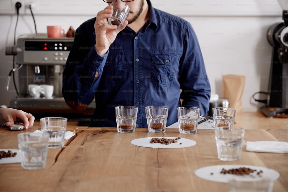 Anonimous portrait of man smelling freshly ground coffee in cups at coffee cupping examine. He is standing near white wall in front of rows with glass cups and coffee machines