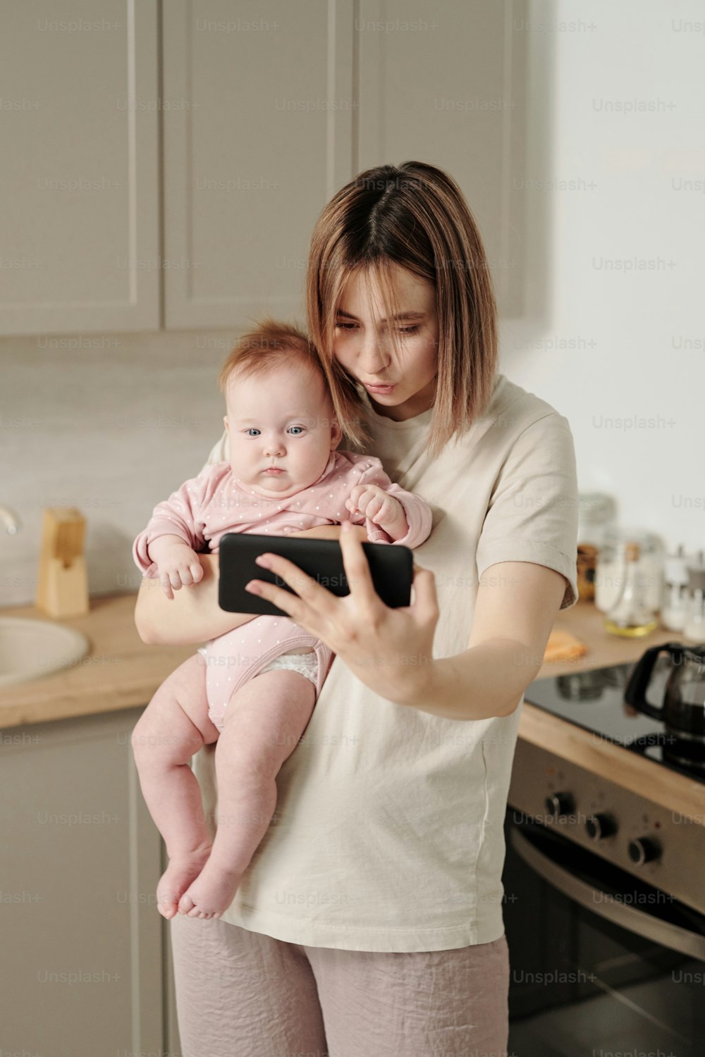 キッチンでビデオチャットでコミュニケーションをとるスマートフォンと赤ちゃんを持つ現代の若い女性
