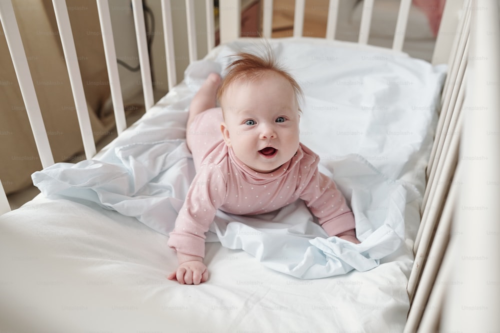 Happy baby girl creeping on white linen in her cradle and looking at camera