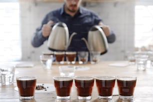 Close up of male barista pouting boiling water into glass cups with ground coffee from two kettles, brewing fresh coffee for tasting examine. Portrait in selective focus