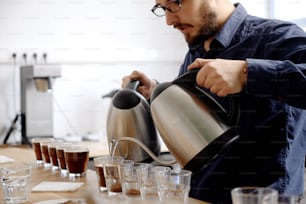 Close up of male barista pouting boiling water into glass cups with ground coffee from two kettles, brewing fresh coffee for cupping exam
