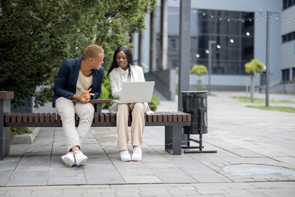 Businessman and businesswoman talking and using laptop while sitting on bench in city. Concept of remote and freelance business work. Idea of business cooperation. Smiling young man and woman