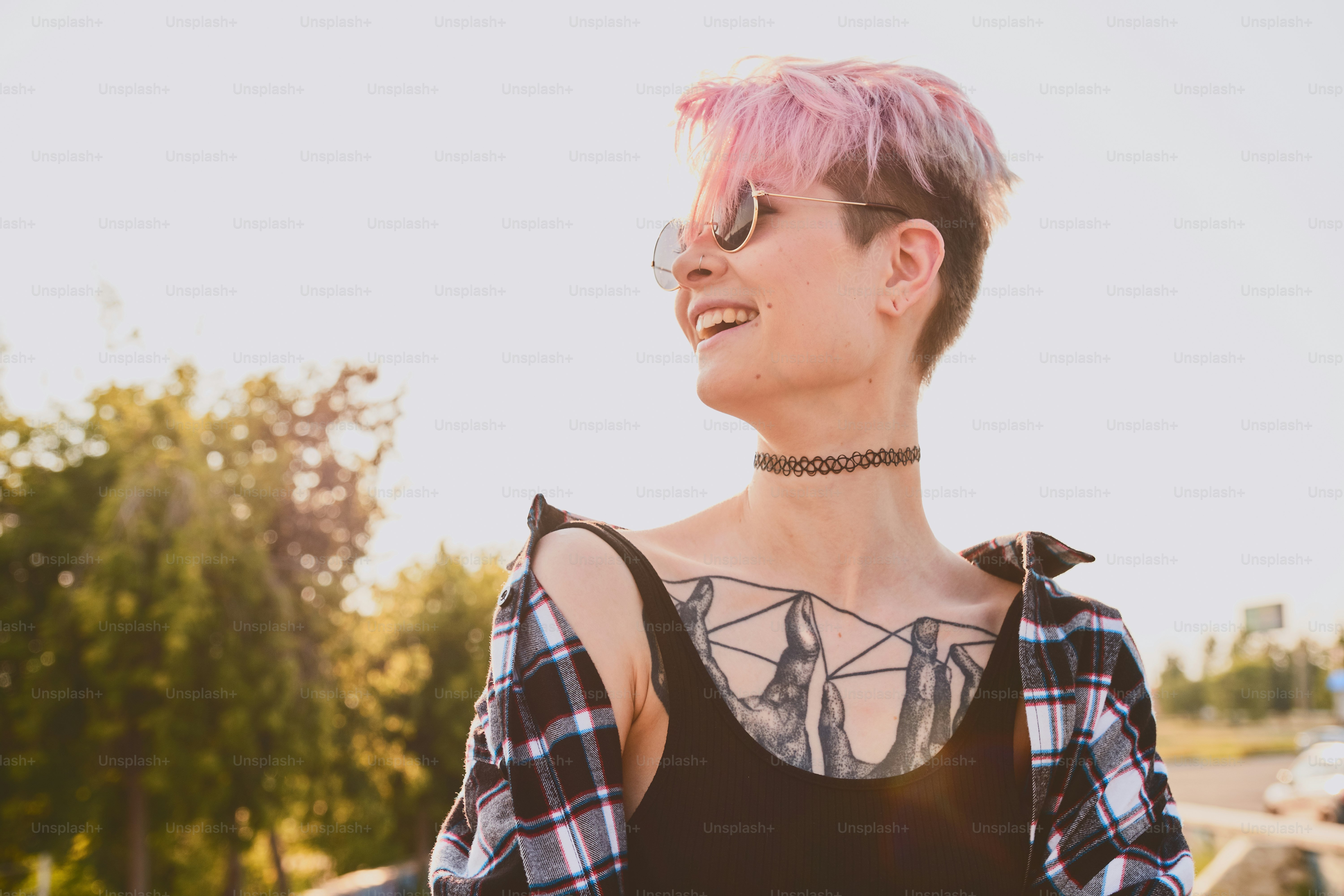 Portrait of daring young tattooed woman with short pink hair, open chest, sitting outdoor in round glasses