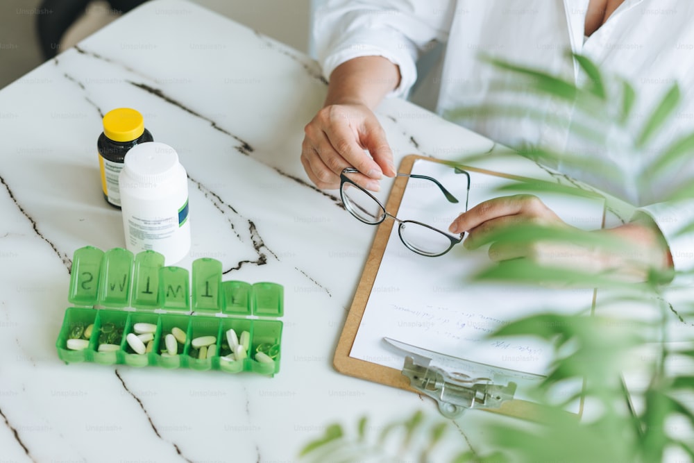 Foto de corte da mulher nutricionista médica plus size em camisa branca trabalhando na mesa com pílulas diárias e notas na sala de escritório moderna brilhante