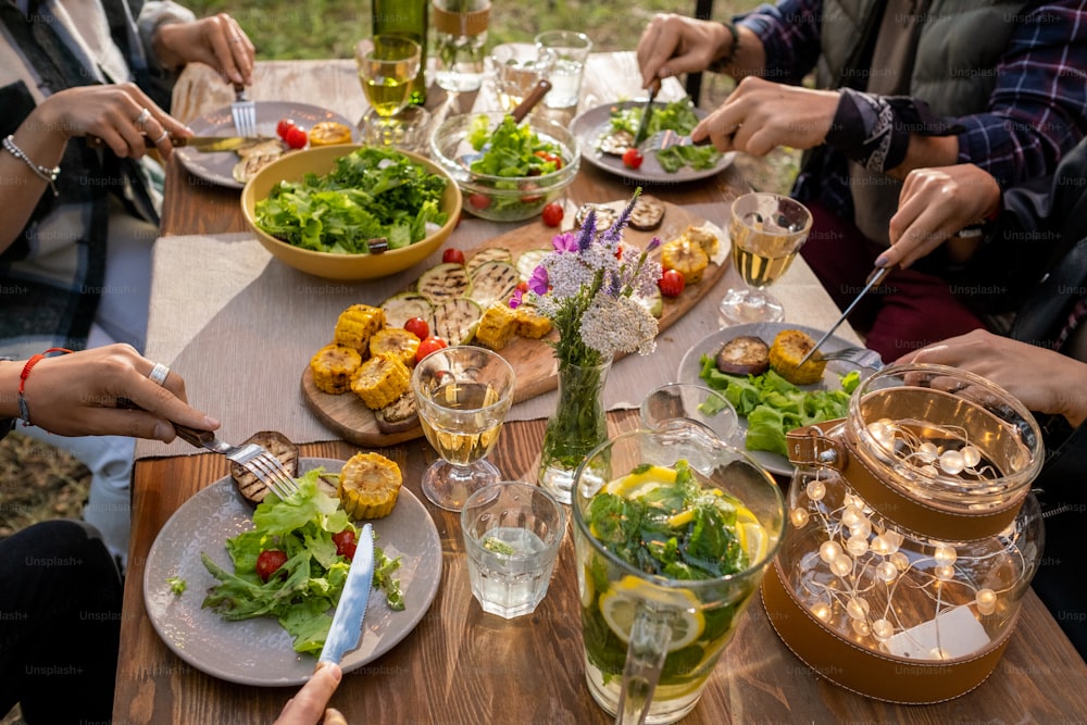 Hands of several friends with knives and forks eating grilled vegetables and salad