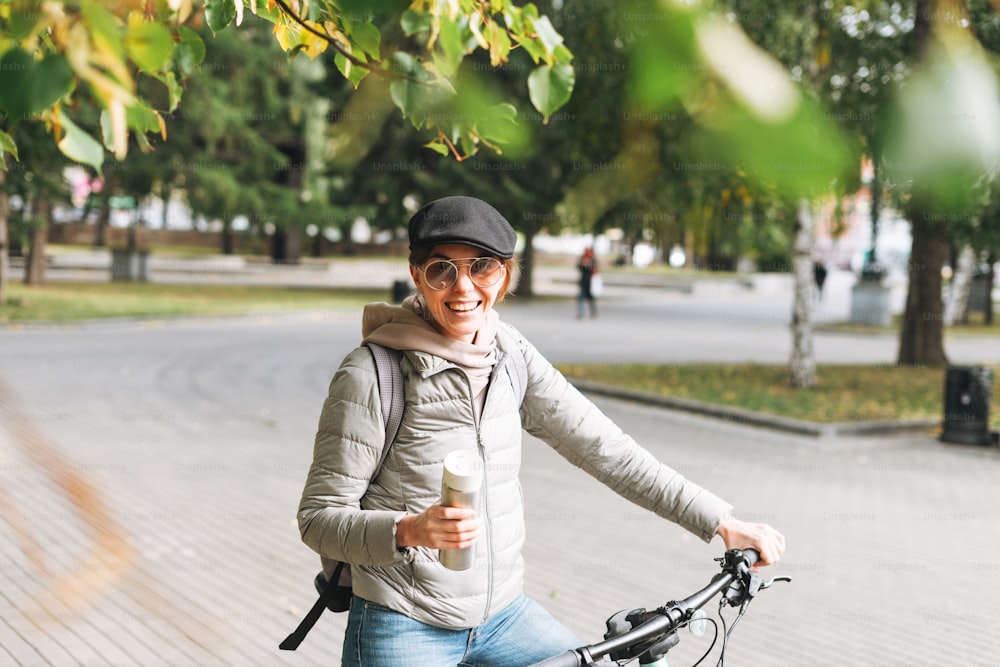 Retrato da mulher bonita jovem da moda no boné e óculos de sol na bicicleta com caneca térmica no dia ensolarado do outono na cidade