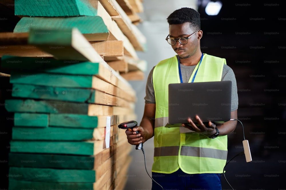 Black warehouse worker using laptop and scanning labels at lumber storage compartment.