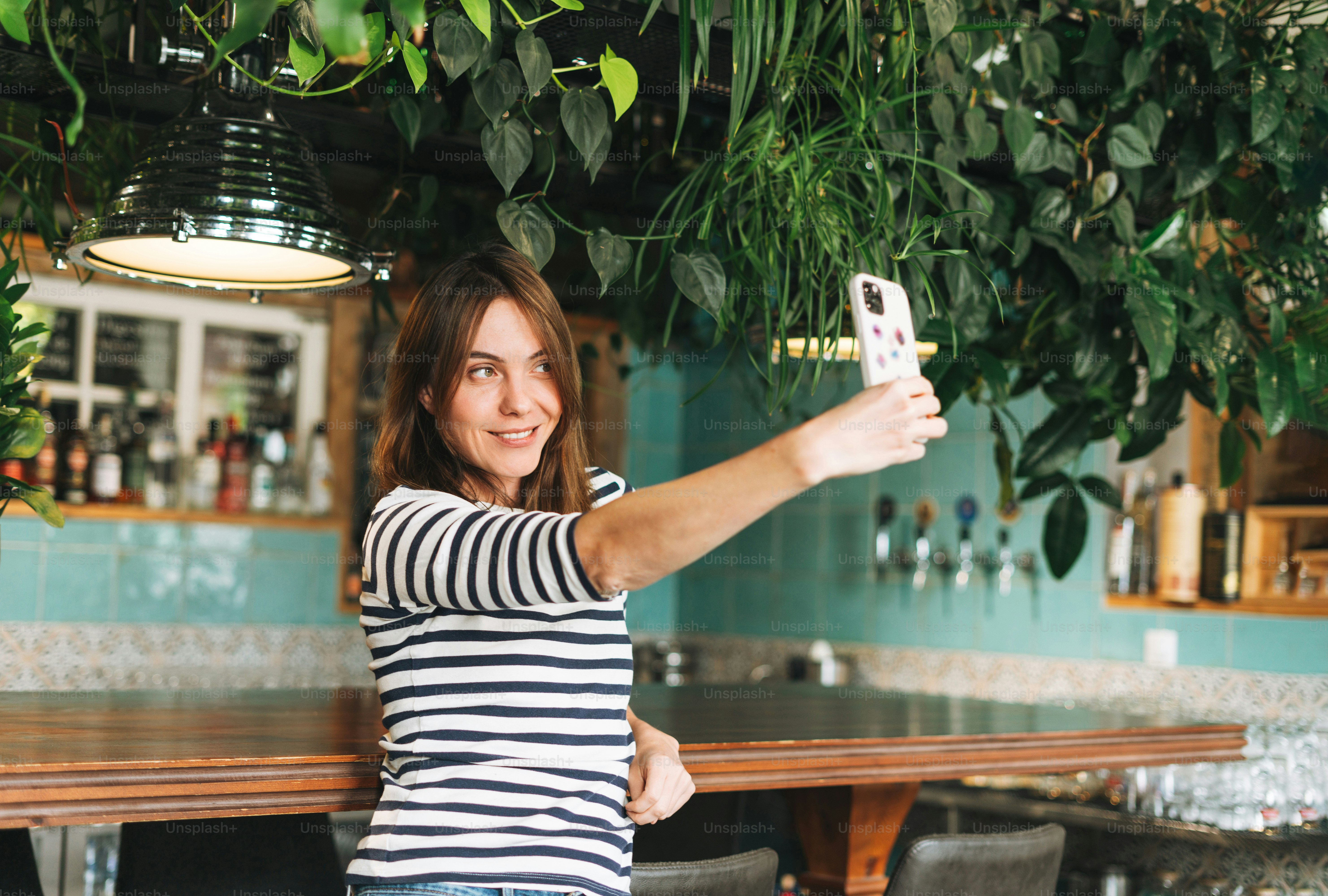 Young attractive smiling woman in casual clothes using social media on mobile phone taking selfie at cafe