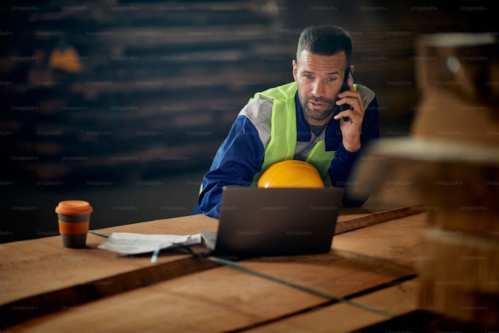 Male worker using laptop and talking on the phone while working and wood distribution warehouse.