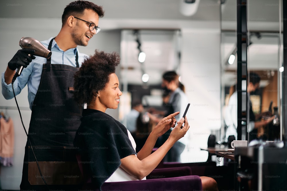 Femme afro-américaine heureuse montrant quelque chose sur son téléphone intelligent à son coiffeur tout en se faisant coiffer au salon.