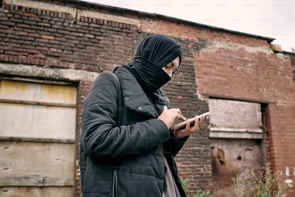 Young refugee female in hijab texting in mobile phone against wall of half ruined building