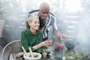 Latin man taking juice for pouring in glass of his european girlfriend during dinner outdoors. Concept of relationship and enjoying time together. Modern domestic lifestyle. Woman sitting at table