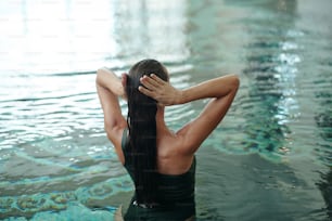 Vista posteriore della ragazza bagnata con i capelli scuri lunghi in piedi nell'acqua della piscina mentre visita il lussuoso centro termale