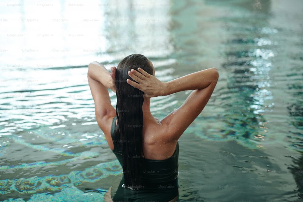 Vue arrière d’une fille mouillée aux longs cheveux noirs debout dans l’eau de la piscine lors de la visite d’un centre de spa luxueux