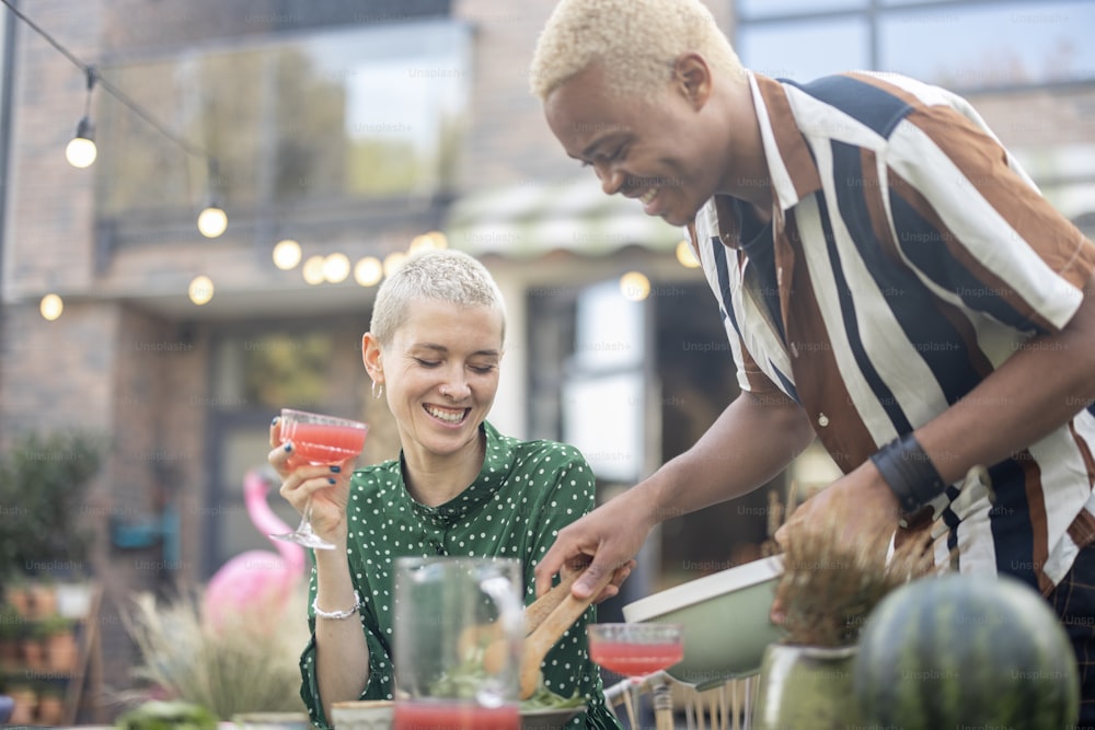 Black man putting salad on plate of his european girlfriend during dinner outdoors. Concept of relationship and enjoying time together. Modern domestic lifestyle. Smiling woman with cocktail