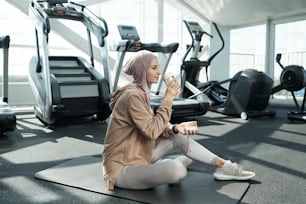 Young serene sportswoman drinking water after sports training while sitting on the floor of gym or fitness center