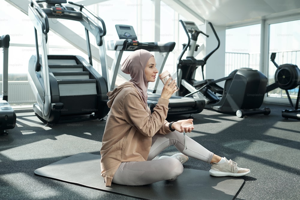 Young serene sportswoman drinking water after sports training while sitting on the floor of gym or fitness center