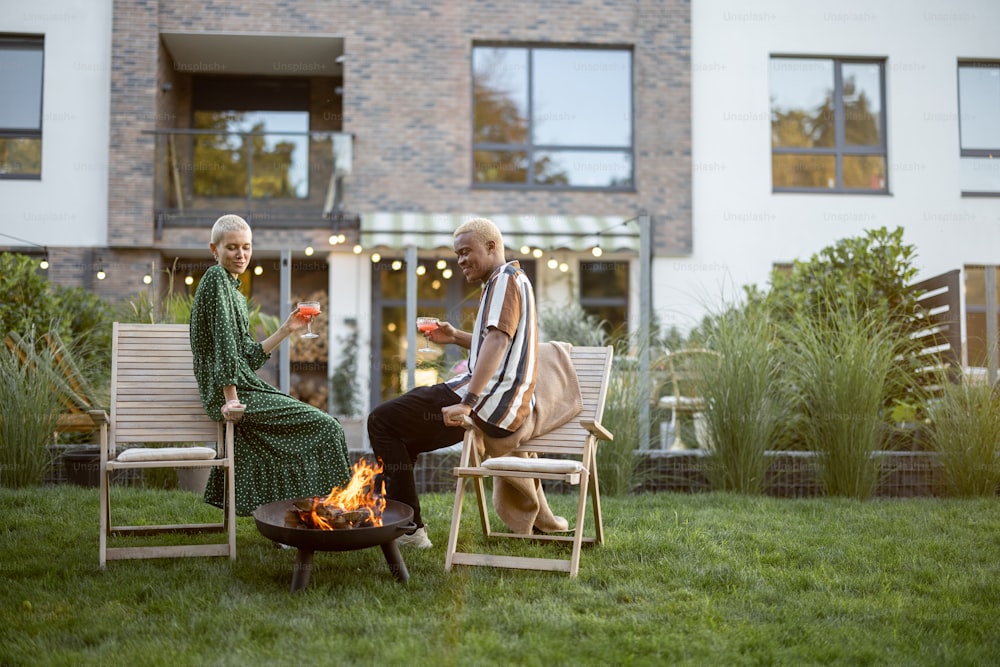 Multiracial couple drinking cocktails during home party in their garden. Burning fire for bbq. Concept of relationship. Modern domestic lifestyle. Black man and european girl enjoying time together
