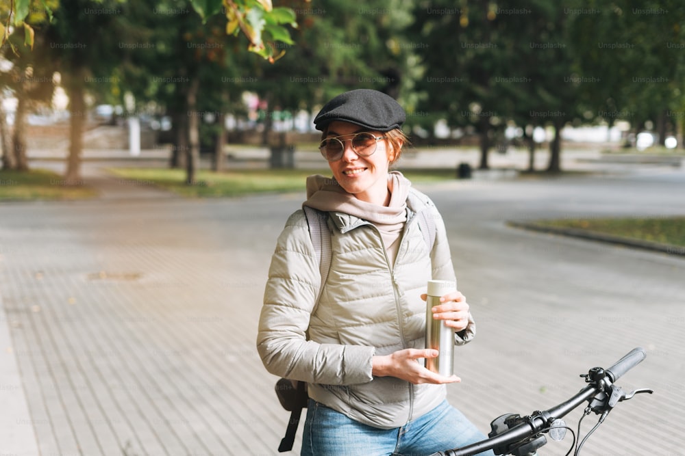 Retrato da mulher bonita jovem da moda no boné e óculos de sol na bicicleta com caneca térmica no dia ensolarado do outono na cidade