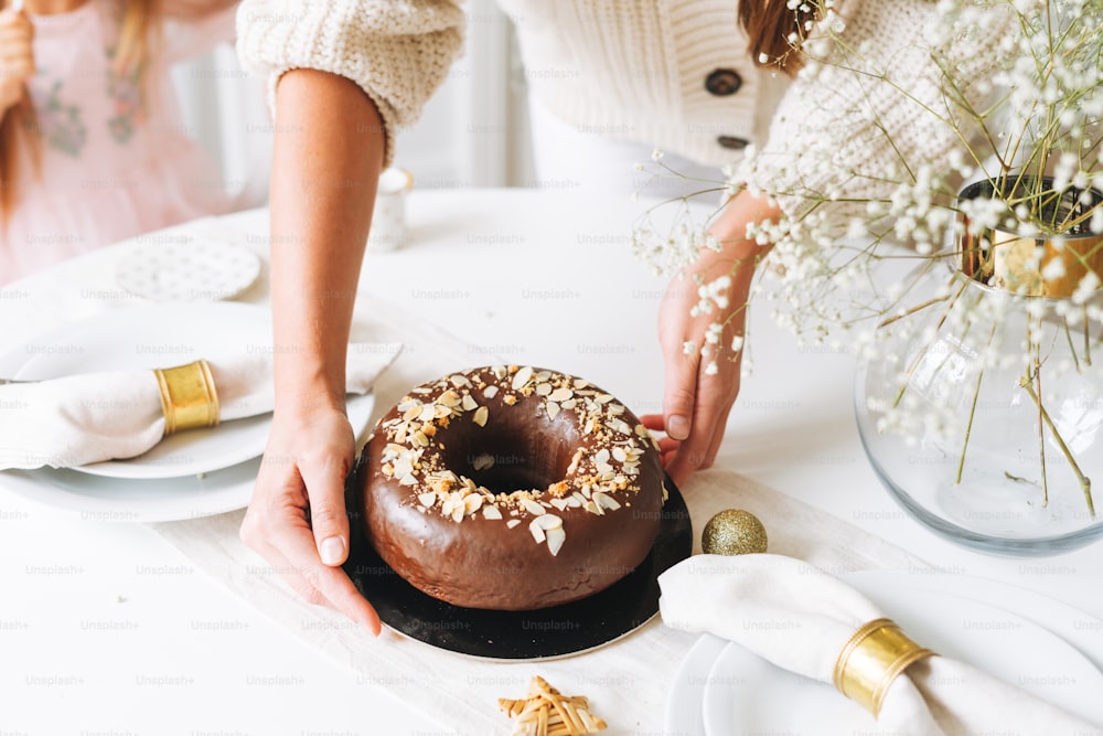 Young woman mother in white holding in hands Large round chocolate almond cake on table with New Year serving, christmas white scandinavian festive table, family event