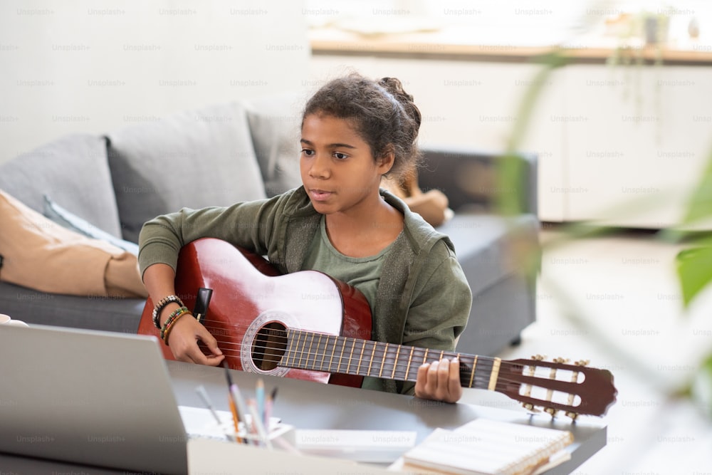 Ernsthafte Schülerin mit Gitarre sitzt am Tisch vor dem Laptop und hört während des Online-Unterrichts auf den Rat des Lehrers