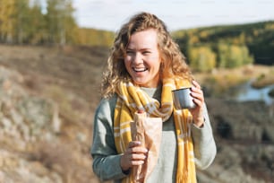 Young happy beautiful woman traveller with curly hair eating hot dog and drinking tea on view background of mountains and river, hiking on autumn nature