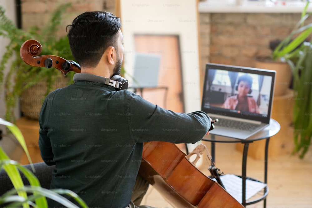 Back view of young music teacher playing cello in front of laptop with cute little schoolboy on screen
