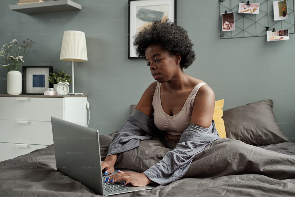 Young female freelancer with laptop sitting in bed after sleep and networking
