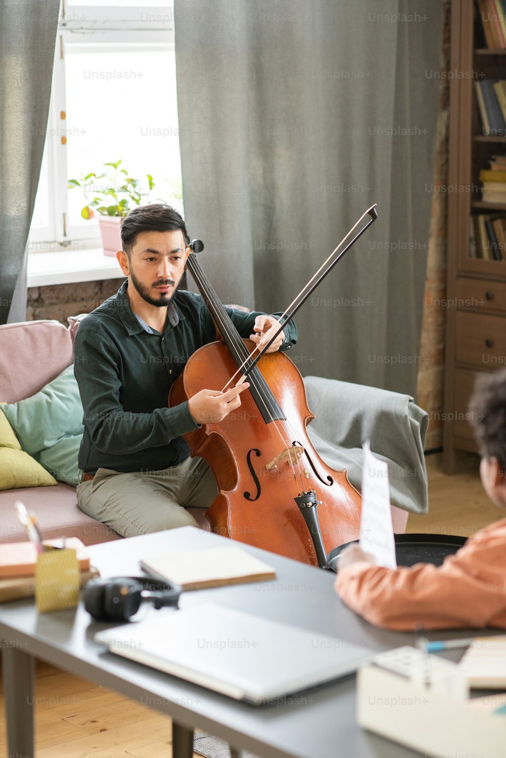 젊은 자신감 있는 음악 교사가 학생 앞에서 소파에 앉아 첼로를 연주하는 방법을 설명합니다.