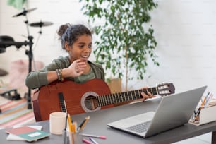 Happy preteen schoolgirl with guitar waving hand to her teacher on laptop screen during online lesson of music