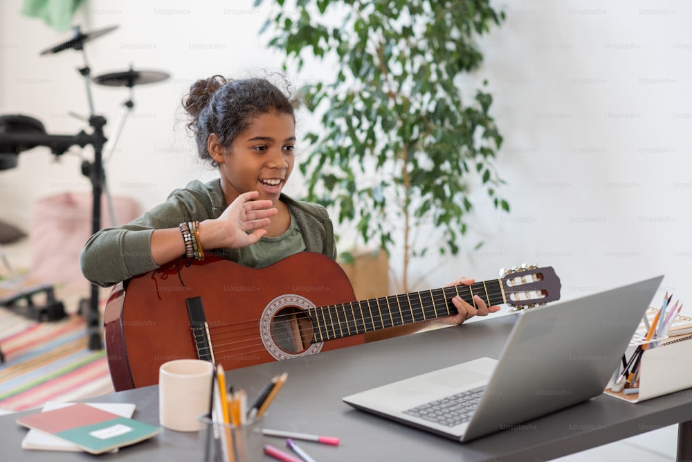 Colegiala preadolescente feliz con la guitarra agitando la mano a su maestro en la pantalla de la computadora portátil durante la lección en línea de música
