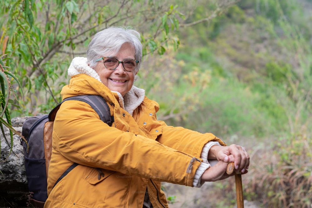 Smiling senior woman outdoor on mountain excursion wearing winter sweater and yellow jacket. Relaxed elderly woman leaning at walking cane looking nature