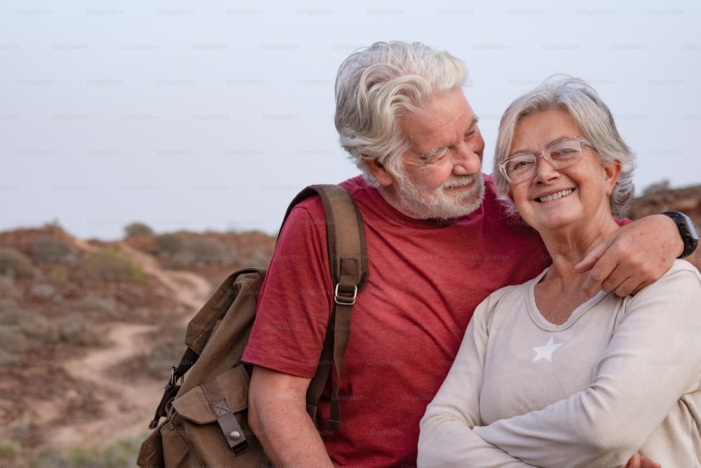 Beautiful senior couple walking in countryside at sunset light, smiling happy. Old mature couple of wife and husband white haired hugging enjoying outdoor and nature