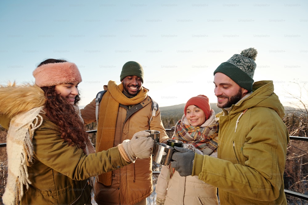 Young multiracial people in winterwear clinking with mugs of hot drinks while enjoying winter weekend