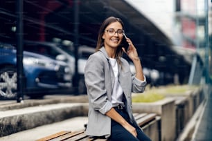 A happy young, elegant woman is sitting outdoors and having a phone call. A woman is sitting on a bench and having a phone call. A businesswoman talking on the phone outside