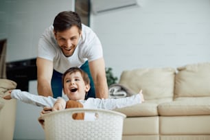 Playful father having fun with his son while pushing him in laundry basket at home.