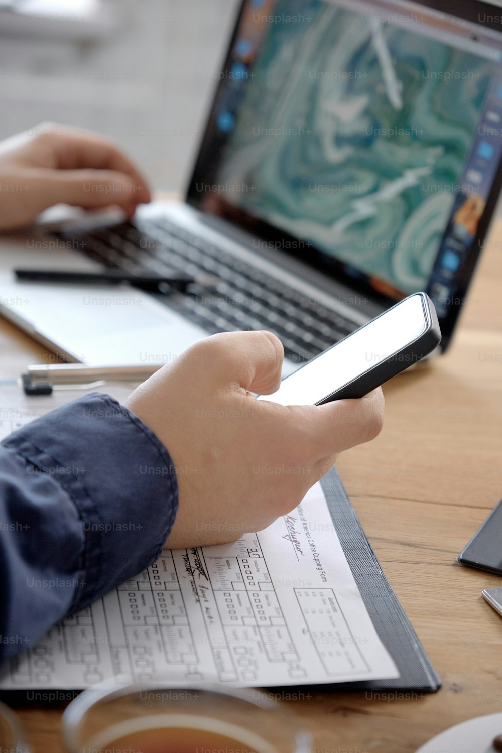 Nahaufnahme der Hände des Mannes mit Smartphone-Bildschirm während der Arbeit am Holztisch mit Papieren, umgeben von Kaffeetassen