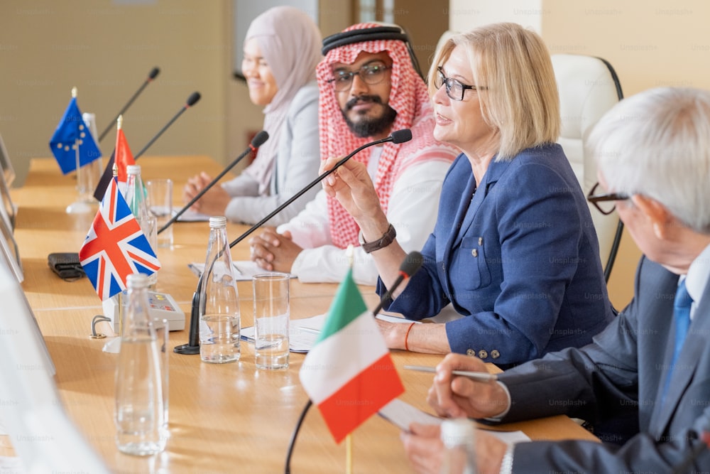 Mature female politician in eyeglasses sitting among multi-ethnic colleagues and speaking into microphone at conference