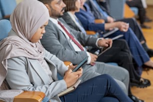 Mujer musulmana concentrada en hiyab usando un teléfono inteligente mientras está sentada en una conferencia de negocios