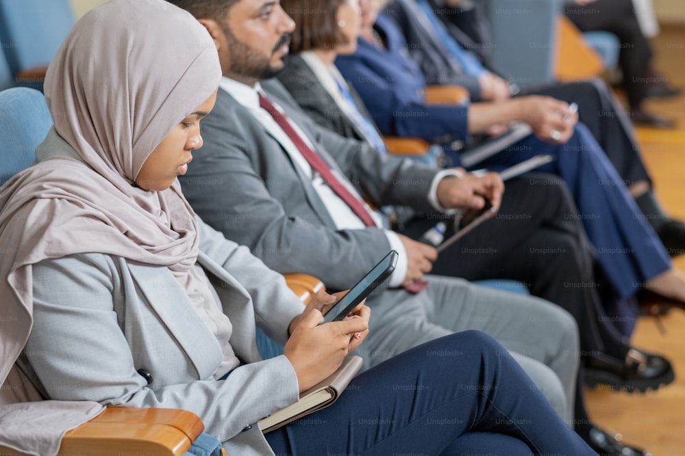 Concentrated Muslim nusinesswoman in hijab using smartphone while sitting at business conference