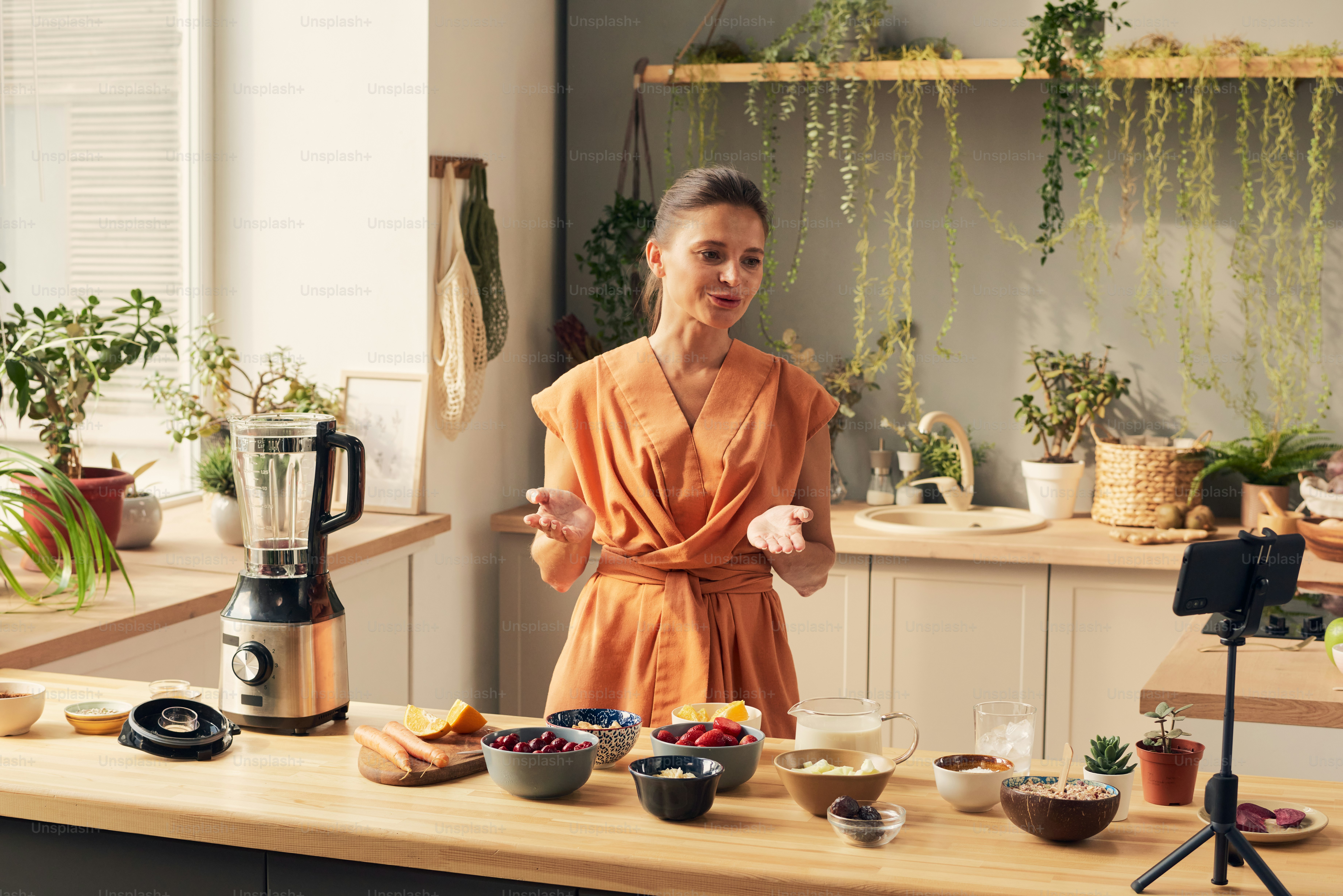 Junge Frau steht am Küchentisch und spricht mit Online-Publikum, während sie vor Smartphone-Kamera Smoothie kocht